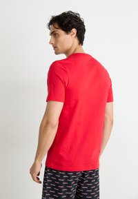 Rotes Baumwoll-T-Shirt mit kurzen Ärmeln und rundem Halsausschnitt. Modell trägt gemusterte schwarze Shorts mit weißem Text.