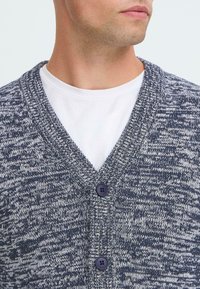 Man som bär en vit t-shirt med rundad hals under en blå och vit stickad knäpptröja.