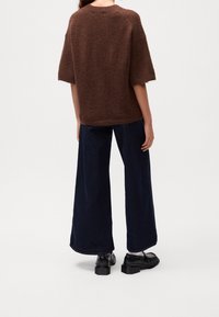 Pull marron en maille texturée avec manches courtes, associé à un pantalon large ample bleu foncé et des chaussures noires à semelle épaisse.