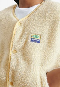 Kremo spalvos vilnonis liemenėlis su minkšta tekstūra, puoštas geltonos spalvos apvadais ir mygtukais. Priekyje yra daugiaspalvė logotipo lygio dėmė.