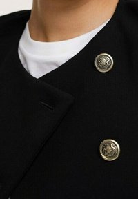 Czarna dwurzędowa marynarka ze złotymi ozdobnymi guzikami. Charakteryzuje się dopasowanym krojem i gładką fakturą materiału. Pod spodem widoczna koszulka.