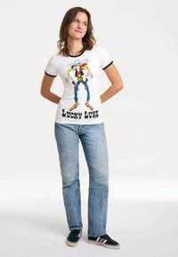 Weißes Baumwoll-T-Shirt mit schwarzem Ringer-Ausschnitt, das eine bunte Lucky Luke-Cartoongrafik und fetten schwarzen Text zeigt, kombiniert mit hellblauen Jeans.
