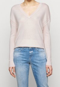 Personne portant un pull léger rose clair à col en V et un jean bleu délavé, debout avec les bras détendus le long du corps devant un fond uni.