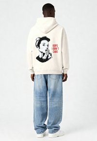 Krémová mikina s kapucňou s grafickou potlačou zobrazujúcou ženu so zakrytými perami prstom a textom "OOPS, NEROBÍ MI TO STAROSTI" v červenej farbe. Bavlnený materiál.