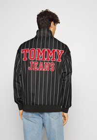 Svart sportjacka med vita smala ränder, med en röd "TOMMY JEANS" logotyp på baksidan. Mjuk tyg, hög krage, elastiska ärmslut och nederkant.