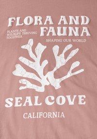Rosa bomulls t-shirt med vit text och ett spräckt korallmönster. Texten lyder "FLORA OCH FAUNA," "SEAL COVE," och "CALIFORNIA."