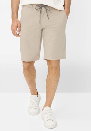Beige knielange shorts met grijze trekkoord, gedragen door een persoon in witte sneakers, staand tegen een effen lichte achtergrond.