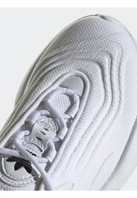 White athletic sneaker with a textured upper, intricate stitching, and distinctive wave patterns. Features white laces and mesh detailing.