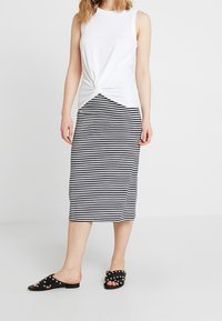 Bílý top bez rukávů s detailem uzlu vpředu, spárován s černo-bílou pruhovanou midi sukni a černými sandály na slip-on se cvočky.
