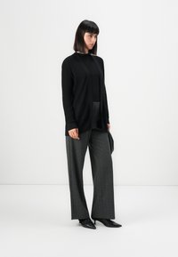 Kvinna med rakt svart hår, iförd svart ribbad cardigan, svart topp, grå vida byxor och svarta spetsiga klackar, stående framför vit bakgrund.