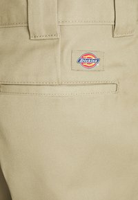 Tissu beige de pantalon Dickies avec poche arrière et passants de ceinture, présentant un écusson rectangulaire de la marque cousu en rouge, jaune et bleu.