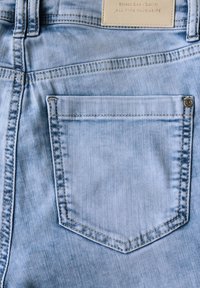 Jean en denim bleu clair avec une poche arrière cousue, des coutures visibles et une étiquette en cuir rectangulaire sur la taille.