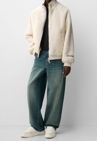 Chaqueta de forro polar color crema con cuello alto, puños acanalados y cierre lateral, combinada con unos vaqueros azules holgados y zapatillas blancas.