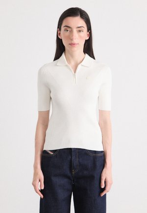 Eine Frau mit langen, dunklen Haaren trägt ein kurzärmeliges, weißes, geripptes Polohemd und dunkle Jeans und steht vor einem schlichten Hintergrund.