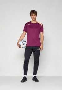 Nike sportoutfit med en lila kortärmad tröja med en orange detalj och svarta figursydda byxor. Modellen håller i en mönstrad fotboll.