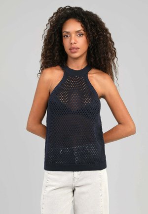 Femme aux cheveux bouclés portant un haut en résille noir sans manches et un pantalon clair, debout les mains derrière le dos sur un fond uni.