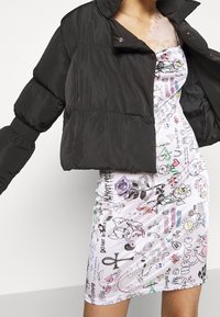 Svart vadderad jacka med quiltat mönster och hög krage, kombinerad med en vit klänning med färgglada klotter och blomster mönster.