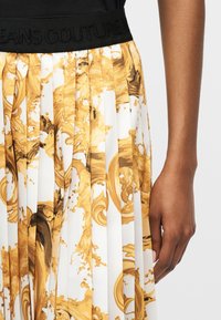 Primer plano de la mano de una persona descansando junto a una falda plisada con estampado dorado de estilo barroco y una cinturilla negra.