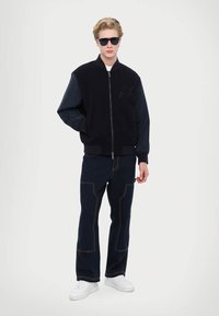 Giacca bomber giovanile in blu navy con maniche nere, chiusura con zip e logo in rilievo. Abbinata a pantaloni scuri con cuciture a contrasto e sneaker bianche.