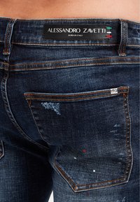 Tmavě modré denimové džíny s opranými detaily, oranžovým šitím a černou štítkem na zadním pásu s nápisem 'Alessandro Zavetti'.