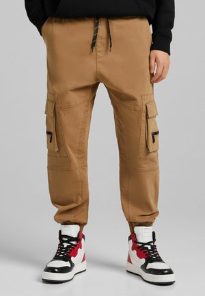 Beige Cargo-Hosen mit elastischem Bund, Seitentaschen und Reißverschlussdetails. Kombiniert mit hochgeschnittenen Sneakers mit roten und schwarzen Akzenten.