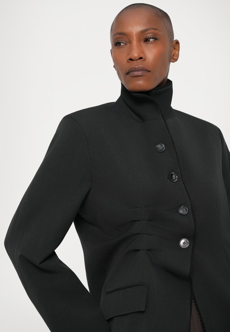 Schwarze, maßgeschneiderte Jacke mit hohem Kragen, die über einen Knopfverschluss, subtile Falten und eine Tasche verfügt. Glattes Material ohne sichtbare Muster.