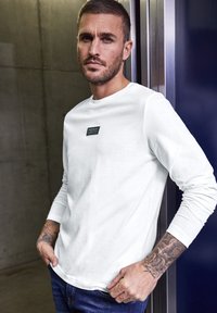 Vit långärmad t-shirt i mjukt tyg med rund hals och en liten svart rektangulär etikett på bröstet. Bärs med blå jeans.