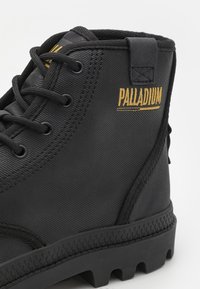 Stivaletto nero con tomaia testurizzata, robusti lacci, colletto imbottito e il marchio giallo "PALLADIUM" vicino al tallone. Suola in gomma con trazione.