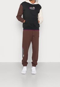 Svart och brun färgblockerad hoodie med vita accenter, som har en framficka och lila branding. Matchas med bruna träningsbyxor som visar logotyp.