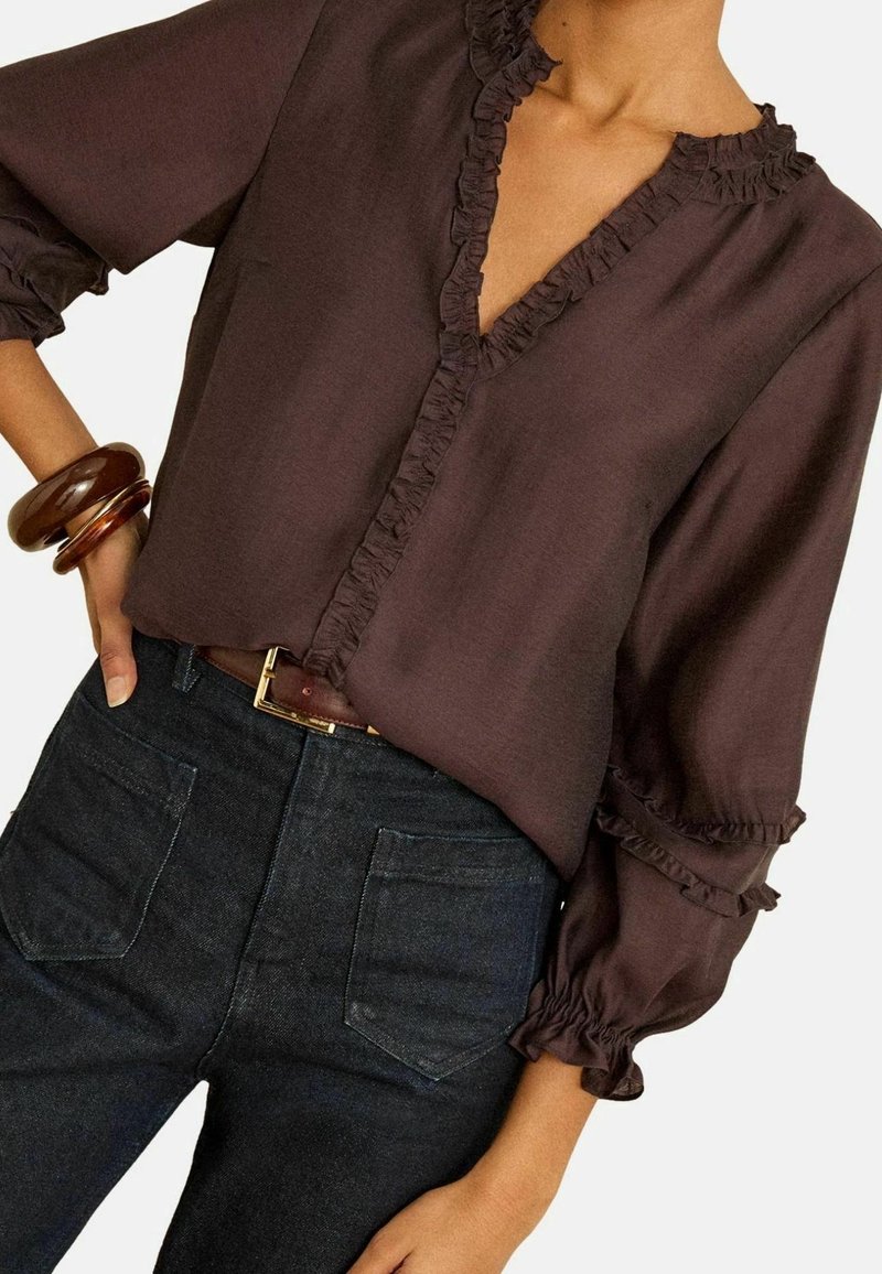 Femme portant un chemisier marron foncé à volants avec manches 3/4, un jean taille haute foncé, une ceinture marron et des bracelets bruns épais au poignet gauche.