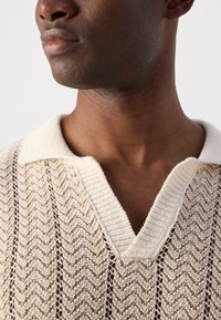 Beige Strick-Polo mit einem strukturierten Chevron-Muster und einem kontrastierenden Rippkragen. Das offene Gewebedesign verbessert die Atmungsaktivität.