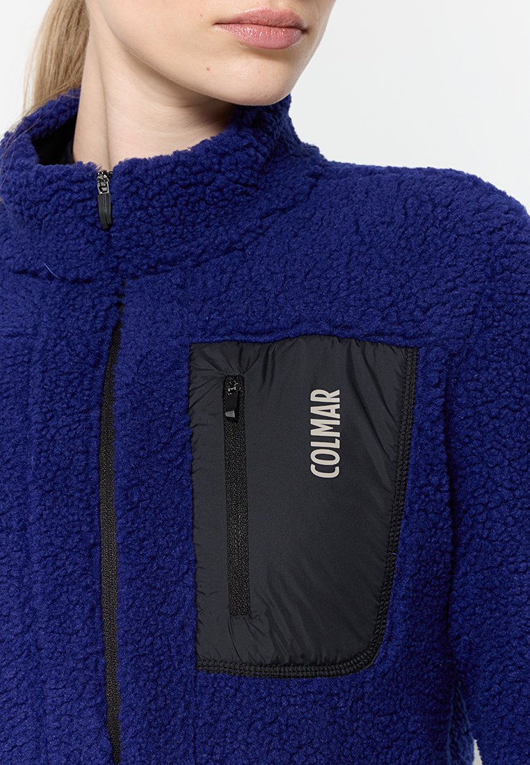 Giacca in fleece blu con colletto alto, dotata di una tasca con zip nera. Texture morbida con un accento di materiale a contrasto.
