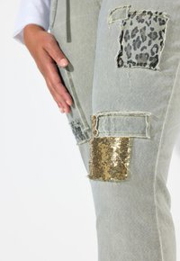 Jeans en denim gris clair présentant de multiples patchs : imprimé léopard, paillettes dorées et zones distendues. Texture lisse avec des détails de couture visibles.