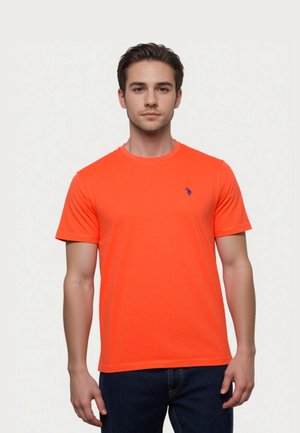 Young man with short brown hair wearing a bright orange short-sleeve t-shirt and dark blue jeans, standing against a plain light background.