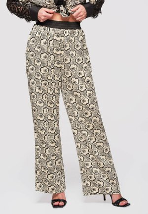 Kobieta ubrana w beżowe spodnie z szerokimi nogawkami ozdobione czarnym kwiatowym wzorem, koszulę związaną w talii oraz czarne sandały na obcasie, stojąca na jednolitym tle.