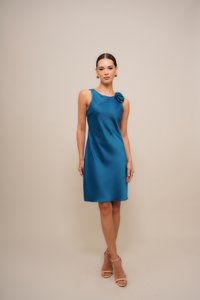 Kvinne som står i en teal satengkjole uten ermer med blomsterdetalj på skulderen og beige ankelrem sandaler mot en enkel beige bakgrunn.