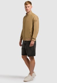 Homme portant une veste beige zippée, un short en jean noir et des baskets blanches, debout devant un fond uni gris clair.