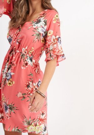 Kvinde iført en koralblomstret kjole med flæseærmer i halv længde og en indsnævret talje, accessoriseret med et tredobbelt armbånd om håndleddet.