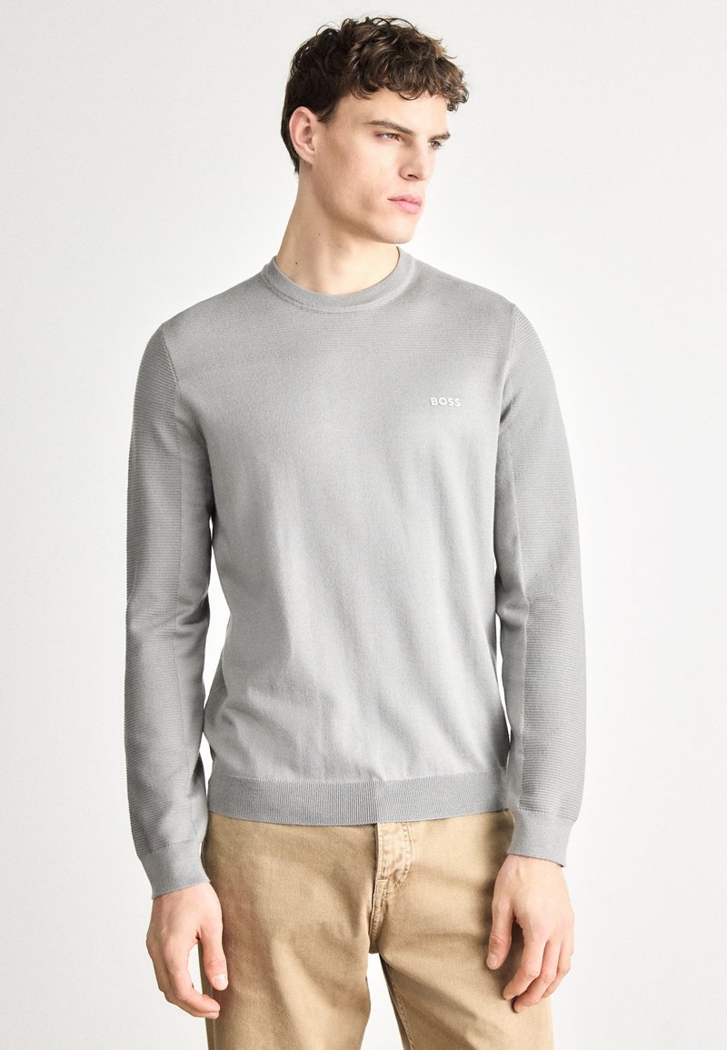 Pulover gri deschis cu guler rotund, mâneci lungi, manșete cu dungi și un logo discret al mărcii pe piept. Textură moale tricotată. Stil casual.