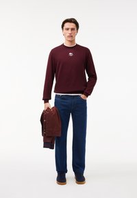 Felpa bordeaux con logo bianco, jeans blu e sneakers navy. Tiene in mano una giacca bordeaux con bottoni a pressione. Texture liscia, vestibilità rilassata.