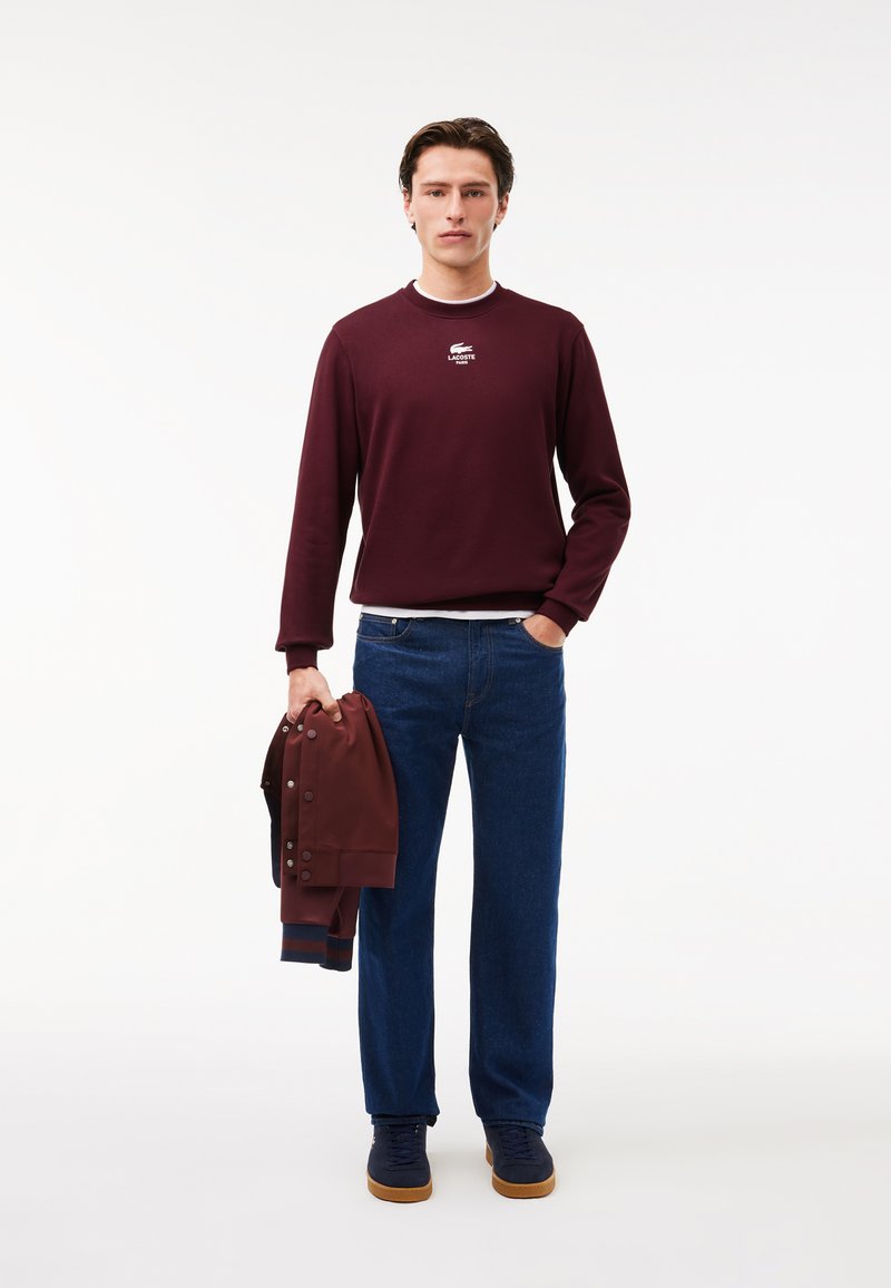 Sweatshirt bordeaux avec logo blanc, jeans bleus et baskets navy. Tient une veste bordeaux avec des boutons-pression. Texture lisse, coupe décontractée.
