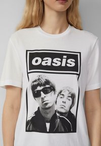 White cotton t-shirt featuring a black and white graphic print of two male figures, with a prominent "oasis" text above the image.