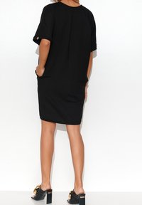 Schwarzes Baumwollkleid mit kurzen Ärmeln und legerem Schnitt, seitlichen Taschen und goldenen Akzentknöpfen an den Ärmeln, kombiniert mit schwarzen Absatzsandalen.