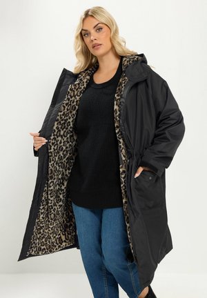 Femme portant un long manteau noir avec une doublure imprimé léopard, un pull noir et un jean bleu, tenant le manteau ouvert pour montrer l’intérieur.