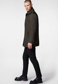 Oliwkowy płaszcz trench z wysokim kołnierzem, z guzikami i dwoma przednimi kieszeniami, zestawiony z wąskimi czarnymi spodniami i masywnymi czarnymi butami.