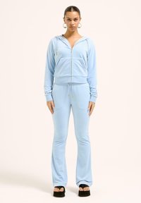 Blå velour zip-up hoodie med framficka, som matchas med flarebyxor. Båda plaggen har dragskoaccents och gyllene detaljer.