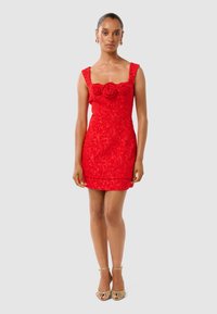 Robe en dentelle rouge avec une coupe ajustée, encolure carrée et trois accents floraux au niveau de la poitrine. Portée avec des sandales à talons hauts dorées et à lanières.
