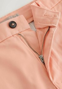 Pantalon de couleur pêche dézippé montrant la fermeture éclair métallique, le bouton, la boutonnière et la ceinture intérieure avec étiquette d'entretien.