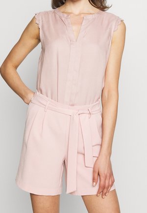 Vrouw draagt een lichtroze mouwloze blouse met kanten rand en een hooggetailleerde bijpassende short met een gebonden stoffen ceintuur.