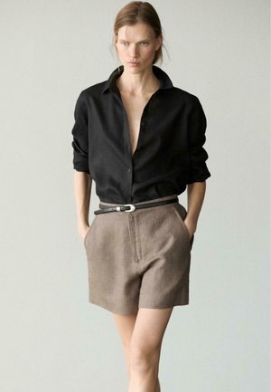 Femme portant une chemise noire à manches longues rentrée dans un short marron taille haute avec une ceinture noire, debout les mains dans les poches.